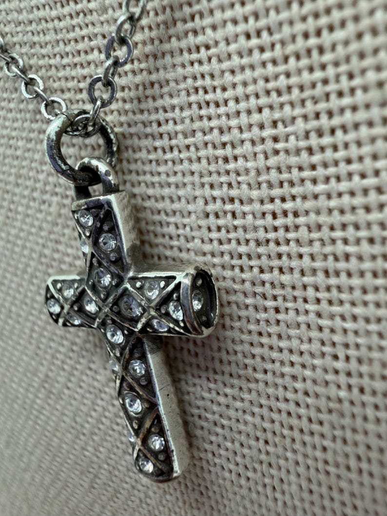 Puede incluir: Collar con colgante de cruz plateada y cadena fina. La cruz est&aacute; adornada con cristales transparentes dispuestos en forma de diamante. El collar se muestra sobre un fondo neutro y texturizado.