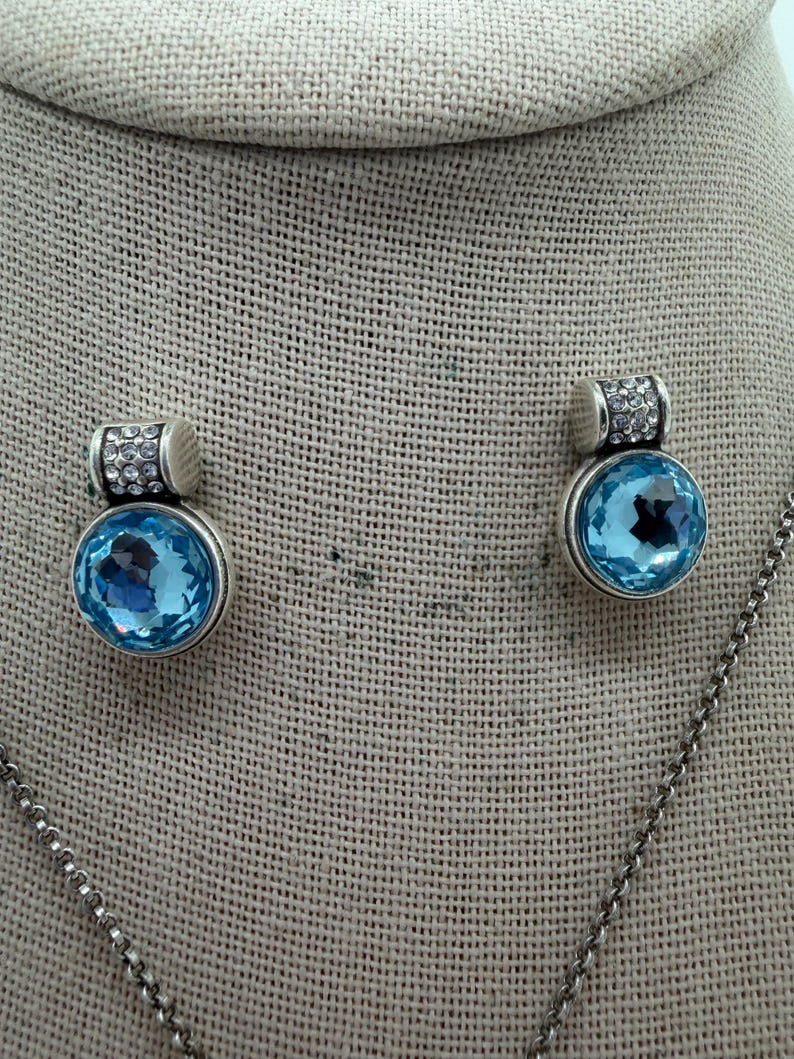 May include: A silver necklace with a pendant and matching earrings. Each piece features a large, round, light blue gemstone. The top of each earring and pendant is decorated with small, clear crystals.