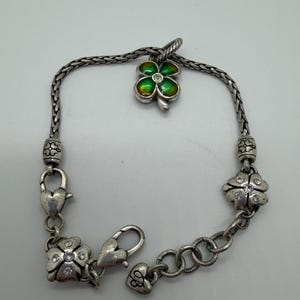 May include: Silver-toned bracelet featuring a four-leaf clover charm with green and gold enamel. The bracelet has clover-shaped accents with clear crystal embellishments and heart-shaped clasps. A delicate piece of jewelry.