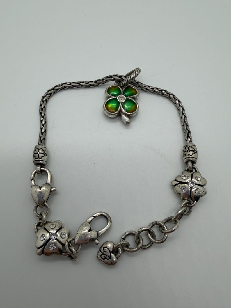 May include: Silver-toned bracelet featuring a four-leaf clover charm with green and gold enamel. The bracelet has clover-shaped accents with clear crystal embellishments and heart-shaped clasps. A delicate piece of jewelry.