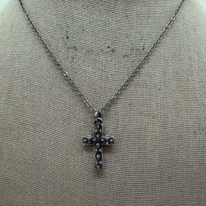 Puede incluir: Un collar plateado con un colgante de cruz. La cruz est&aacute; adornada con peque&ntilde;as piedras preciosas transparentes. El collar se muestra sobre un fondo de tela de color neutro, resaltando el dise&ntilde;o de la joya. La cadena es delicada.