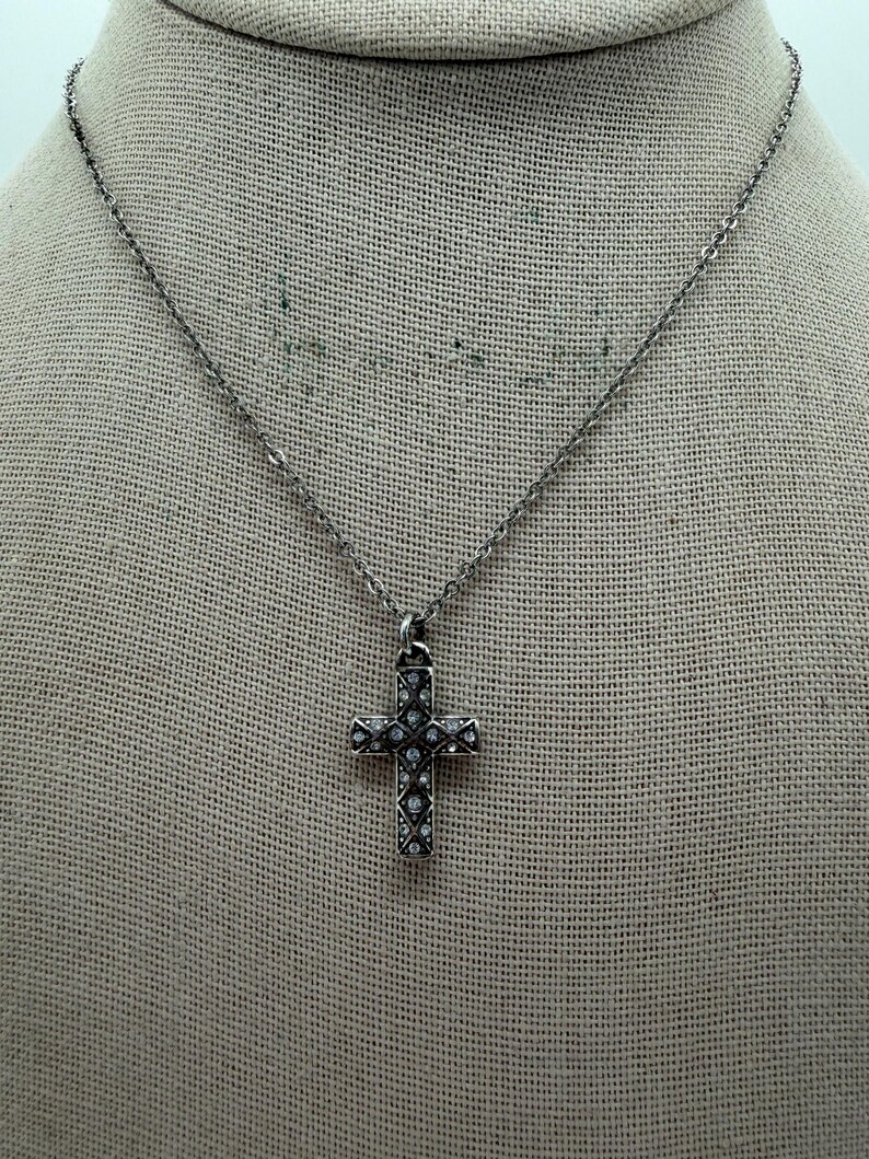 Puede incluir: Un collar plateado con un colgante de cruz. La cruz est&aacute; adornada con peque&ntilde;as piedras preciosas transparentes. El collar se muestra sobre un fondo de tela de color neutro, resaltando el dise&ntilde;o de la joya. La cadena es delicada.