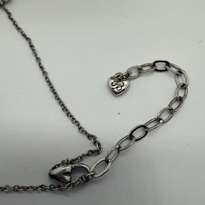 Puede incluir: Collar plateado con un colgante de cruz y un dije en forma de coraz&oacute;n. La cadena es delicada y el dije de coraz&oacute;n tiene un dise&ntilde;o grabado. El collar est&aacute; sobre una superficie blanca.