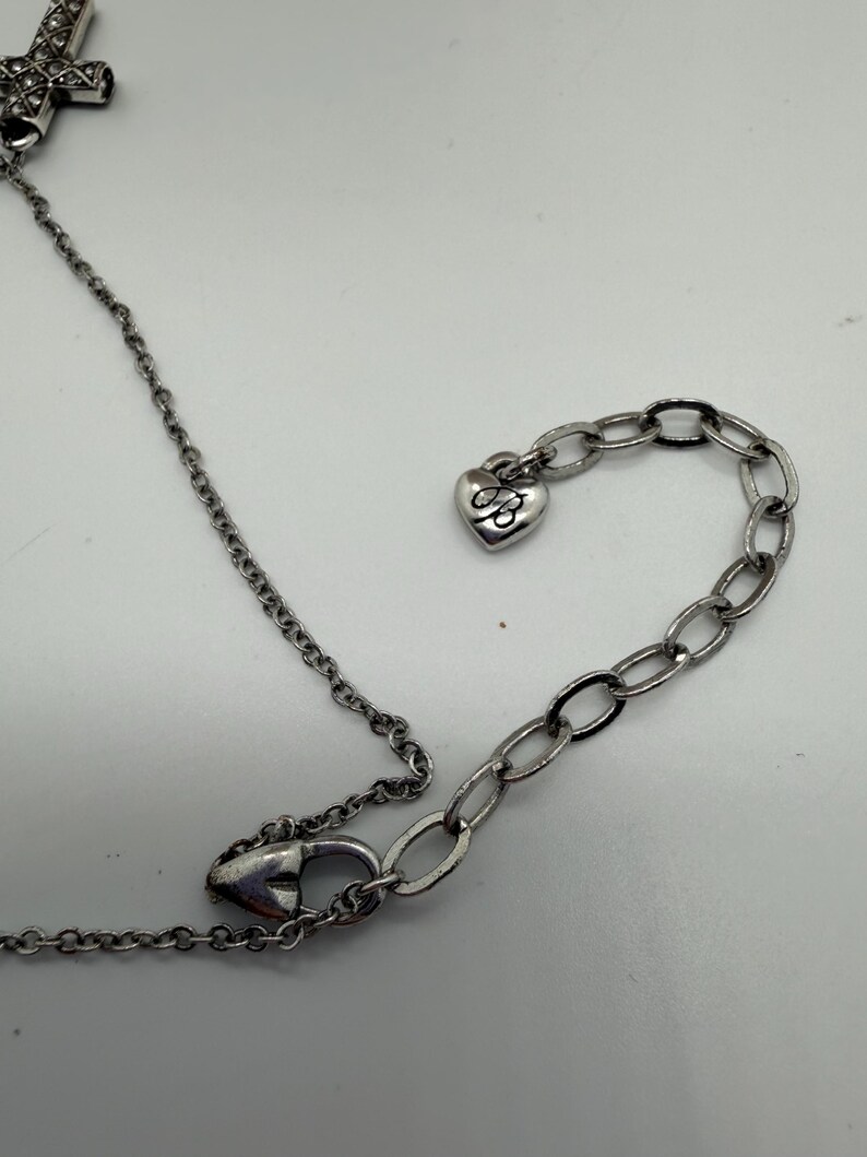 Puede incluir: Collar plateado con un colgante de cruz y un dije en forma de coraz&oacute;n. La cadena es delicada y el dije de coraz&oacute;n tiene un dise&ntilde;o grabado. El collar est&aacute; sobre una superficie blanca.