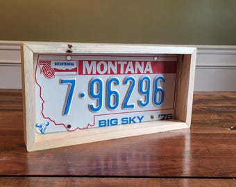 Puede incluir: Una placa de matrícula vintage de Montana enmarcada en un marco de madera natural. La placa es blanca con letras azules y rojas. La placa dice "Montana" "7-96296" "Bicentenario" "Big Sky" "'76".