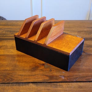 Puede incluir: Un organizador de madera con cuatro ranuras, teñido de un color marrón intenso. El organizador tiene una base negra y es ideal para guardar objetos pequeños como bolígrafos, lápices u otros artículos de oficina.