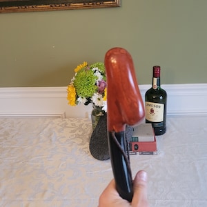 May include: A wooden walking stick with a brown and black handle. The handle is carved with a unique shape and has a leather strap. The stick is being held by a hand.