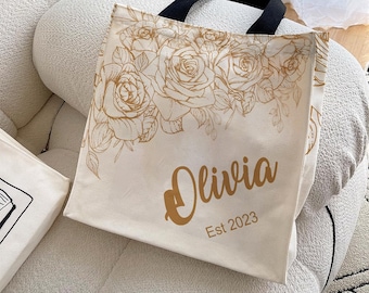 Könnte beinhalten: Cremefarbene Tragetasche mit schwarzem Griff. Sie hat ein goldenes Rosenblumendesign und den Namen "Olivia" mit "Est 2023" in Gold aufgedruckt. Die Tasche liegt auf einem cremefarbenen Stuhl.