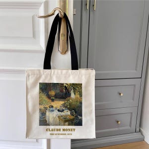 May include: A white canvas tote bag with black handles. The bag features a painting of Claude Monet's "The Luncheon" with the artist's name and the year 1873 printed in gold below the image.