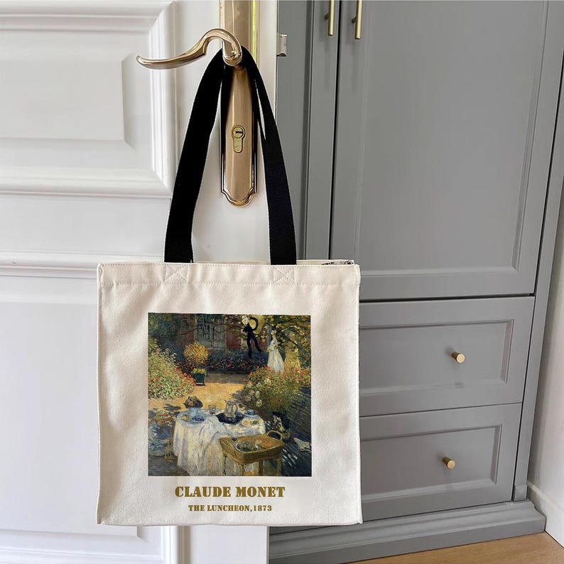May include: A white canvas tote bag with black handles. The bag features a painting of Claude Monet's "The Luncheon" with the artist's name and the year 1873 printed in gold below the image.