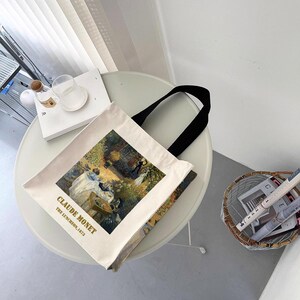 May include: A white canvas tote bag with black handles. The bag features a painting of Claude Monet's "The Luncheon on the Grass" with the artist's name and the date of the painting printed below the image.