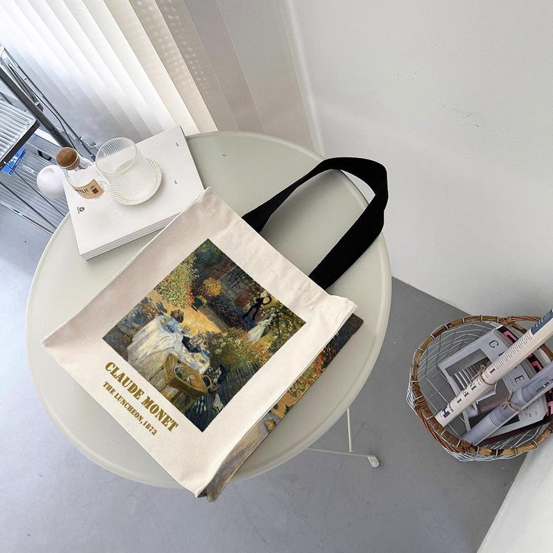 May include: A white canvas tote bag with black handles. The bag features a painting of Claude Monet's "The Luncheon on the Grass" with the artist's name and the date of the painting printed below the image.