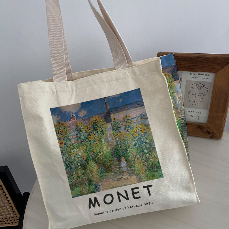 May include: A white canvas tote bag with a printed image of a painting by Claude Monet. The painting depicts a garden with sunflowers and a path leading up to a house. The text "MONET" and "Monet's garden at V&eacute;theuil, 1880" is printed below the image.