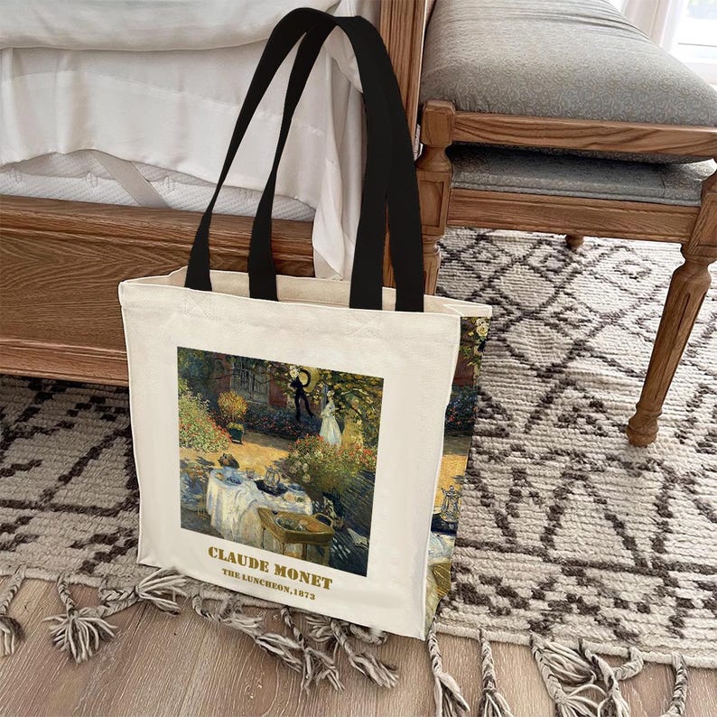 May include: A white canvas tote bag with black handles. The bag features a printed image of Claude Monet's painting "The Luncheon, 1873".