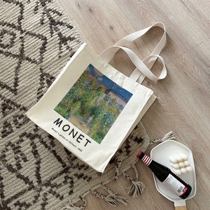 May include: A white canvas tote bag with a printed image of a painting by Claude Monet. The painting depicts a garden with sunflowers and a blue sky. The text "MONET" and "Monet's garden at V&eacute;theuil, 1880" is printed on the bag.
