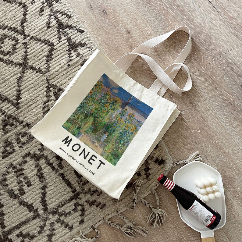 May include: A white canvas tote bag with a printed image of a painting by Claude Monet. The painting depicts a garden with sunflowers and a blue sky. The text "MONET" and "Monet's garden at V&eacute;theuil, 1880" is printed on the bag.