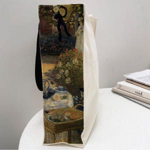 May include: A white canvas tote bag with black handles. The bag features a printed image of a painting depicting a garden scene with people, flowers, and a table setting. The painting is in a style reminiscent of Impressionism.