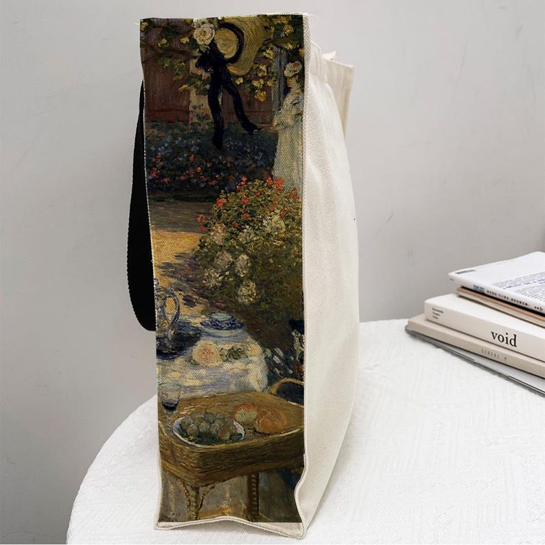 May include: A white canvas tote bag with black handles. The bag features a printed image of a painting depicting a garden scene with people, flowers, and a table setting. The painting is in a style reminiscent of Impressionism.