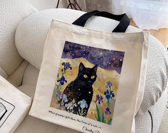 Puede incluir: Una bolsa de lona blanca con asas negras presenta un gato negro sentado en un campo de flores moradas y azules. El fondo es un cielo nocturno estrellado. La cita "What greater gift than the love of a cat... - Charles Dickens" está impresa en la bolsa.