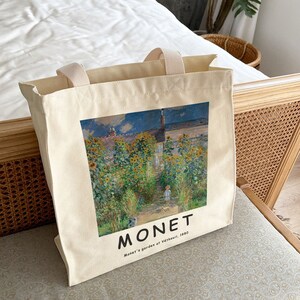 May include: A white canvas tote bag with a printed image of a painting by Claude Monet. The painting depicts a garden with sunflowers and a path leading to a house. The text "MONET" and "Monet's garden at V&eacute;theuil, 1880" is printed below the image.