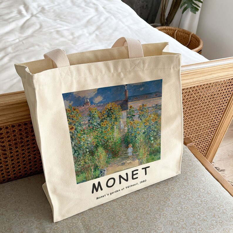 May include: A white canvas tote bag with a printed image of a painting by Claude Monet. The painting depicts a garden with sunflowers and a path leading to a house. The text "MONET" and "Monet's garden at V&eacute;theuil, 1880" is printed below the image.