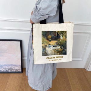 May include: A white canvas tote bag with a black handle. The bag features a painting of a garden scene with people having a picnic. The text "Claude Monet, The Luncheon, 1873" is printed in gold on the bag.