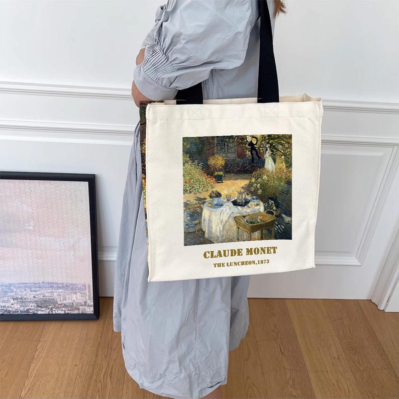 May include: A white canvas tote bag with a black handle. The bag features a painting of a garden scene with people having a picnic. The text "Claude Monet, The Luncheon, 1873" is printed in gold on the bag.