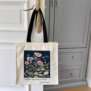 Może przedstawiać: Biała torba tote z płótna z czarnymi uchwytami. Torba ma niebieskie tło z kwiatowym wzorem i cytat Claude'a Moneta: "Muszę mieć kwiaty, zawsze i zawsze."