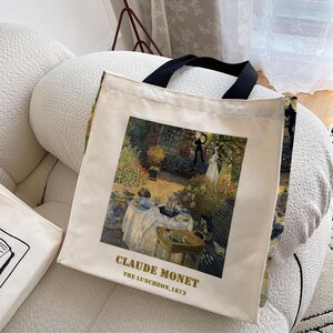 May include: A white canvas tote bag with black handles. The bag features a painting of a garden scene with people having a picnic. The text "Claude Monet" and "The Luncheon, 1873" is printed on the bag.