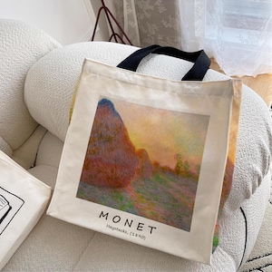 Può includere: Una borsa tote in tela bianca con manici neri. La borsa presenta un dipinto di covoni di fieno in un campo al tramonto di Claude Monet. Il testo "MONET Haystacks, (1890)" è stampato sulla borsa.