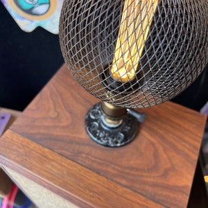 May include: A decorative table lamp with a spherical wire cage shade. The lamp features a vintage-style Edison bulb, casting a warm glow. The lamp is mounted on a dark wooden base, creating a rustic and industrial aesthetic.