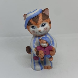 Pode incluir: Uma figurinha de cerâmica de um gato vestindo um pijama listrado azul e branco e um chapéu azul, segurando uma figurinha de gato menor vestindo uma camisa rosa e um chapéu azul.
