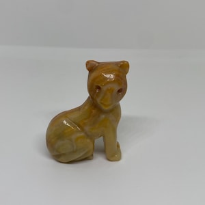 Peut inclure: Petite figurine sculptée représentant un ours assis. L'ours est un mélange de couleurs marron clair et beige, avec une finition brillante. La figurine présente des détails, notamment de petites oreilles et un visage défini.