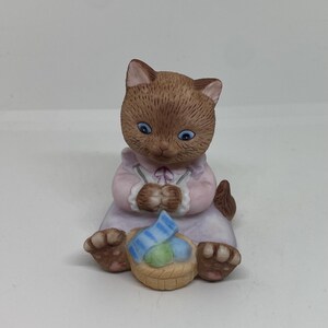 May include: A brown and white porcelain figurine of a cat wearing a pink dress and holding a basket of blue and white striped eggs.