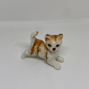 Pode incluir: Uma pequena figura de porcelana de um gato branco e marrom tigrado com olhos azuis. O gato está em uma pose brincalhona com o rabo levantado.