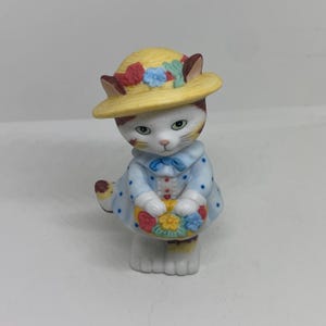 Pode incluir: Uma figurinha de porcelana branca de um gato que usa um chapéu de palha amarelo com flores e um vestido azul com bolinhas brancas. O gato está segurando uma cesta de flores.