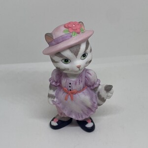 Pode incluir: Uma pequena figura de porcelana de um gato usando um chapéu rosa com uma flor e um vestido roxo. O gato tem listras cinzas e brancas e olhos verdes.