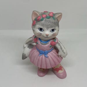Pode incluir: Uma figurinha de porcelana de um gato cinza e branco vestindo um vestido rosa com um laço azul e uma coroa de flores. O gato está usando sapatos de balé rosa.