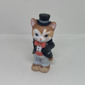 Pode incluir: Uma figurinha de cerâmica de um gato vestindo um smoking preto, uma gravata borboleta vermelha e um chapéu-coco preto. O gato tem pêlo castanho e branco e olhos azuis.