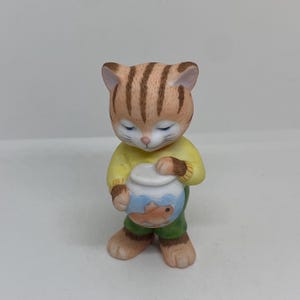 Pode incluir: Uma figurinha de cerâmica de um gato segurando um aquário com um peixe dourado dentro. O gato está vestindo um suéter amarelo e calças verdes.
