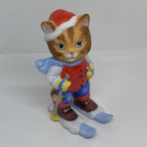 Pode incluir: Uma figurinha de cerâmica de um gato com um chapéu de Papai Noel vermelho e branco, uma echarpe azul, uma jaqueta vermelha, calças azuis e botas de esqui marrons. O gato está segurando bastões de esqui e está em pé em esquis.