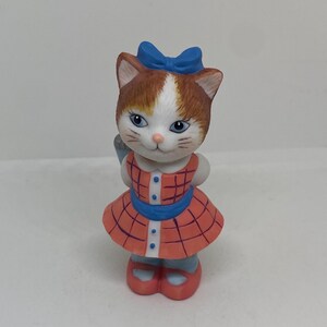 May include: A porcelain figurine of a cat wearing a red and white plaid dress with a blue sash and a blue bow. The cat has brown and white fur and blue eyes.