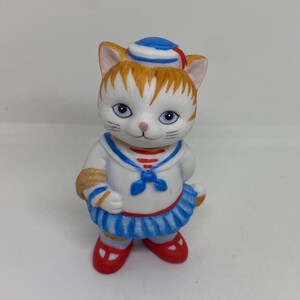 Pode incluir: Uma figurinha de porcelana branca de um gato vestido de marinheiro. O gato está usando um chapéu de marinheiro azul e branco, uma camisa de marinheiro branca com listras vermelhas e azuis e uma saia azul e branca. O gato está segurando uma corda em sua pata direita.