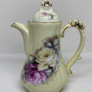 May include: A vintage porcelain coffee pot with a lid, featuring a pale yellow base color and a floral design of purple and cream roses. The handle and spout are accented with gold trim, and the lid has a gold floral finial.