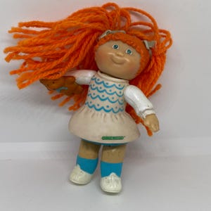 May include: A small plastic Cabbage Patch Kid doll with orange yarn hair, wearing a white dress with blue and white wavy pattern, blue socks, and white shoes. The doll is holding a brown teddy bear.