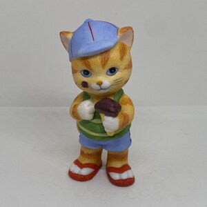 Peut inclure: Une figurine en céramique d'un chat portant une casquette de baseball bleue, une chemise rayée verte et blanche, un short bleu et des sandales rouges. Le chat tient un cornet de glace au chocolat.
