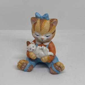 Pode incluir: Uma figura de cerâmica de um gato tigrado marrom e laranja vestindo um macacão azul e um laço azul, segurando um gatinho branco.