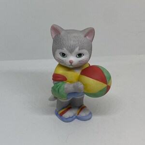 Pode incluir: Uma figura de gato cinza e branco vestindo uma camisa amarela, vermelha e verde e sandálias azuis. O gato está segurando uma bola de praia vermelha, amarela e verde.