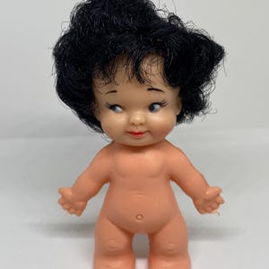 May include: A small, nude-colored plastic doll with black rooted hair. The doll has a painted face with blue eyes and a closed-mouth smile.