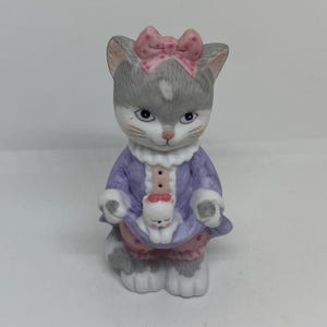 Op de afbeelding: Een keramiek beeldje van een grijze kat die een paarse jurk met roze stippen en een roze strik draagt. De kat houdt een kleiner wit kitten vast.